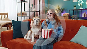 Girl child in 3D glasses eating popcorn watching TV serial sport game film with beagle dog at home