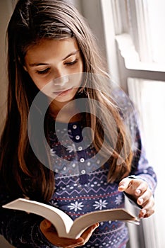 Girl, child and book for reading in home with thinking, learning and language by window for studying. Kid, literacy and