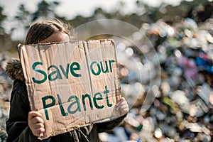 Girl child with cardboard sign with text \'Save our planet\' in front of blurry mountain of garbage