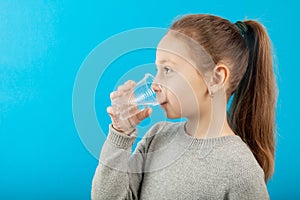 Girl child drinks pure mineral water from glass, portrait on blue background