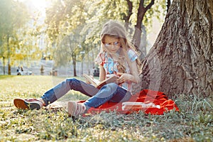Girl child in glasses reading book in the park, on the grass near the tree