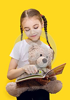 Girl child reads a book and holds a teddy bear on a yellow background