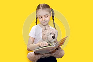 Girl child reads a book and holds a teddy bear on a yellow background