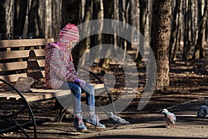 A girl child in a red jacket, hat and glasses feeds doves in the park with seeds.