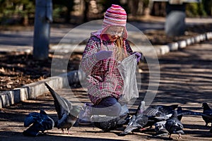 A girl child in a red jacket, hat and glasses feeds doves in the park with seeds.