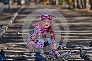 A girl child in a red jacket, hat and glasses feeds doves in the park with seeds.