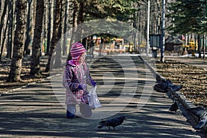 A girl child in a red jacket, hat and glasses feeds doves in the park with seeds.