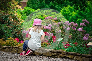 Girl child relaxing in  flower garden rear view. Adorable little kid sitting around growing flowery. Young caucasian female child