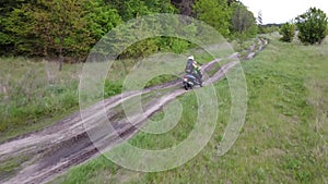 A girl and a child riding a moto scooter or motorcycle along a dirt road between field and forest.Aerial, drone footage.