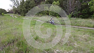 A girl and a child riding a moto scooter or motorcycle along a dirt road between field and forest.Aerial, drone footage.