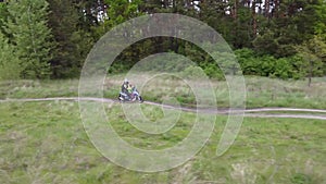 A girl and a child riding a moto scooter or motorcycle along a dirt road between field and forest.Aerial, drone footage.