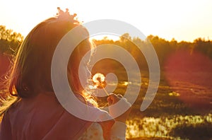 girl child in the twilight light of the setting sun, holding a white fluffy dandelion, glare of sun rays   .