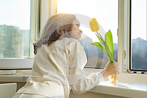 Girl child with yellow tulip flower, near the window sill looks out the window opening glass, sunny spring day