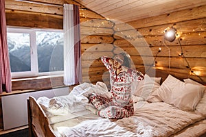 Girl in Christmas pajamas relaxing in bed inside cozy log cabin with winter view