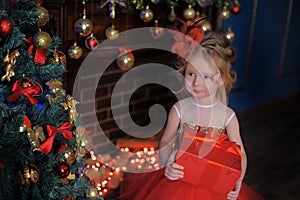 Girl at the Christmas tree in a red elegant dress with a box with gifts in her hands