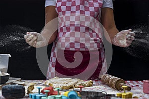 Girl claps hands and spraying flour over dough on dark background. Professional girl baker  is preparing cookies