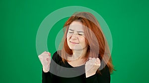 The girl clenches her fists, closes her eyes, makes a wish, black clothes, red hair, chromakey green background.