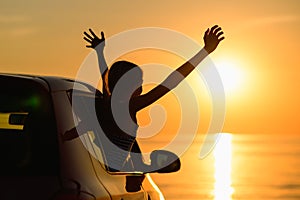 The girl climbed out of the car window with two hands up and looks at the beautiful sunset.