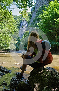 Girl with climbing helmet and a backpack sits on a rock at Varghis gorge, Persani mountains, Romania, near the river.