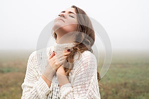 Girl closed her eyes, praying in a field during beautiful fog. Hands folded in prayer concept for faith