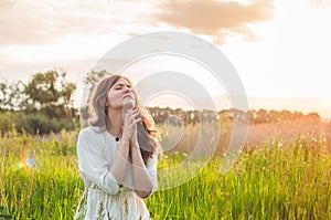 Girl closed her eyes, praying in a field during beautiful sunset. Hands folded in prayer concept for faith