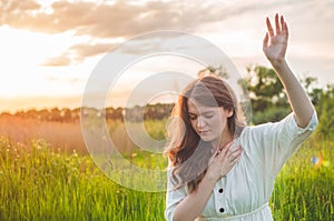 Girl closed her eyes, praying in a field during beautiful sunset. Hands folded in prayer concept for faith