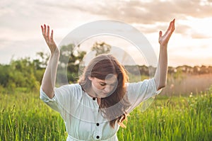 Girl closed her eyes, praying in a field during beautiful sunset. Hands folded in prayer concept for faith