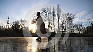 Girl in coat and figure skates sliding on ice at sunset time. Young woman skating on frozen river at frozen day. Shining