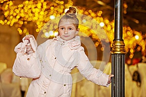 Girl in coat stands, leaning one hand on lamp post