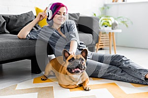 Girl with colorful hair and headphones sitting on floor and petting french bulldog