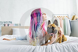 Girl with colorful hair petting cute french bulldog and looking away while laying