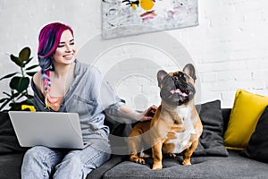 Girl with colorful hair sitting on sofa, using laptop and petting french bulldog