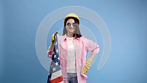 A girl in a construction helmet and gloves holding american flag stands on a blue background