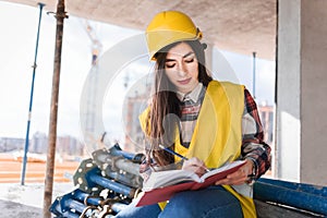 Girl in a construction helmet and vest writes in a notebook at a construction site