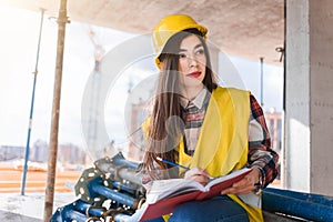 Girl in a construction helmet and vest writes in a notebook at a construction site