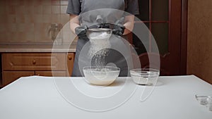 The girl cook in black gloves and a gray apron sifting the flour into a glass bowl with dough