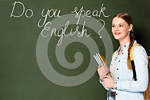 Girl with copybooks and backpack standing near chalkboard with do you speak English lettering