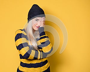 Girl covering face with hand. Shame, model wearing woolen cap and sweater, isolated on yellow background. Ashamed young woman