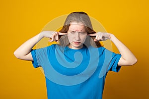 Girl covering her ears with hands for not to hear anything. Photo of young girl dressed in casual clothes on yellow background