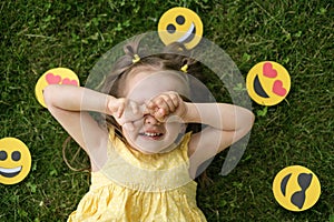The girl covers face with her hands, lying on the grass on the lawn, surrounded by various icons of emoticons