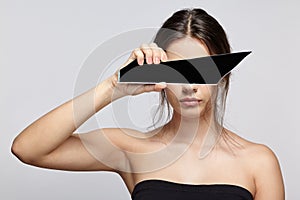 Girl covers her face with a shard of the mirror. Female with mirror shard in hand posing on gray background
