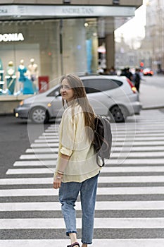 Girl crosses the road at the pedestrian crossing looking over her shoulder. Teenager against the backdrop of busy street