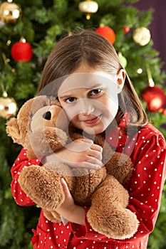 Girl Cuddling Bear In Front Of Christmas Tree