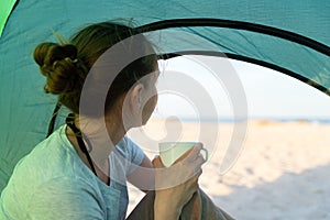 Girl with cup in hands sits in tent and looks at beach and sea. Camping. adventure lifestyle