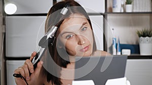 Girl curls her hair. Attractive woman in bathroom wrapped in towel looks in mirror, holds curling iron in her hands and makes