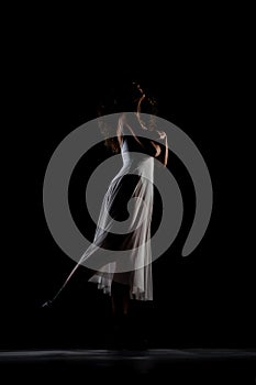 Girl with curly hair making ballet poses. Side lit silhouette of ballerina in white dress and black boots against black background
