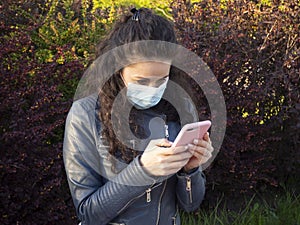 A girl with curly hair in a medical mask is looking in her smartphone in the green spring park