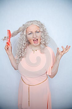 Girl with curly white hair can`t comb her loose dry hair and is upset about it. a blonde woman stands in a pink dress with an oran