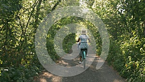 Girl cyclist is riding retro bike in green park in sunlight. Woman in hat rides old bicycle on forest road at sunrise. Happy femal