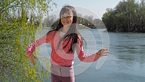 The girl dances by the water. Child on a sunny day in the fresh air. The wind develops the hair. A little girl is dancing on the r